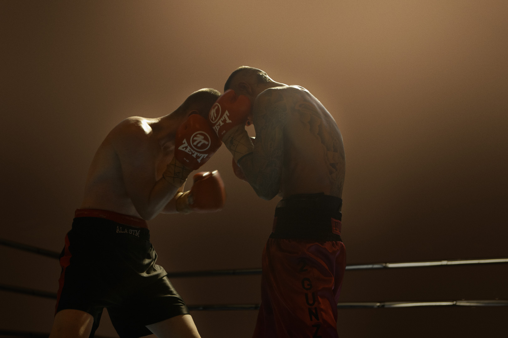 Two boxers in red gloves face off in a boxing ring under bright lights, one with tattoos on his back, during an intense match.