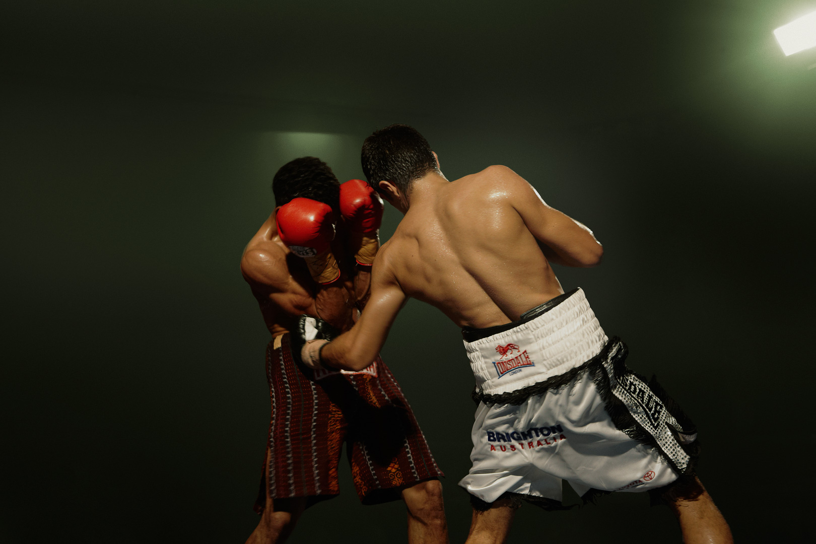 Two male boxers compete in a boxing ring, wearing red boxing gloves, patterned shorts, and athletic shoes in an indoor setting with bright lighting.