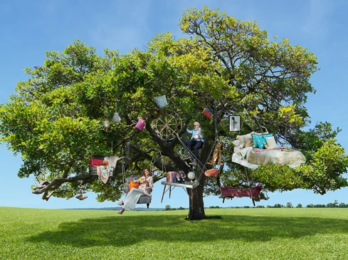 Surreal scene of furniture and people in a large tree against a blue sky over a green field. Woman sits on a couch and another climbs ladder.