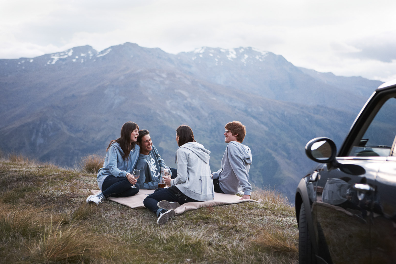 Group of friends enjoys a mountainside picnic with a car parked nearby, creating a picturesque scene of outdoor leisure and adventure.
