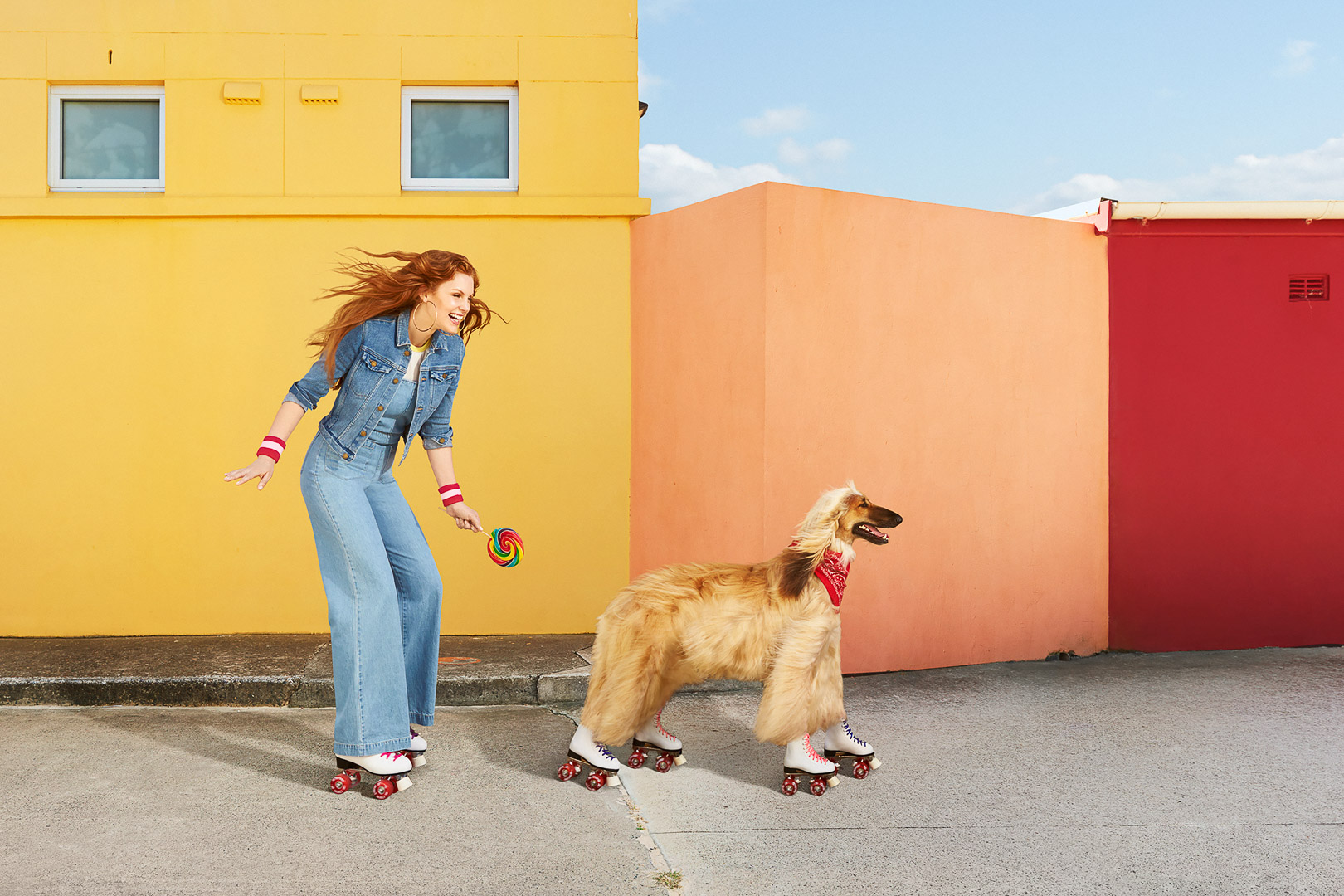 A red-haired woman and Afghan Hound dog roller skate on pavement by bright yellow, orange, and red blocky building walls outdoors.