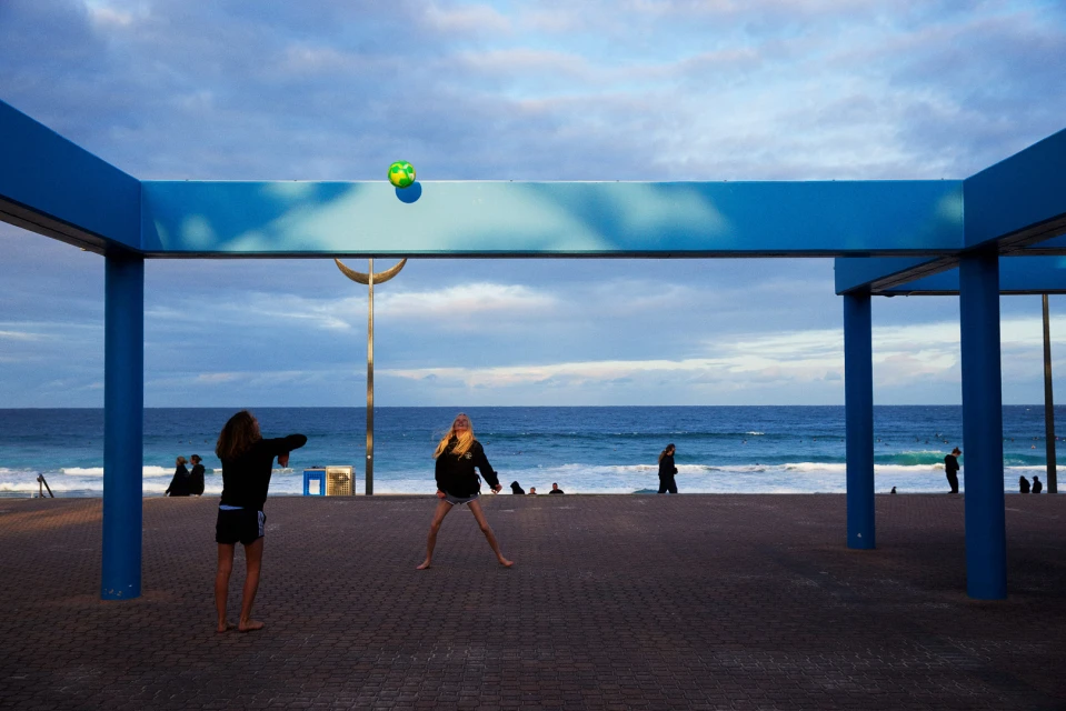 Two young women play with a soccer ball on a paved promenade by the sea under a blue-columned structure, beachgoers in background.