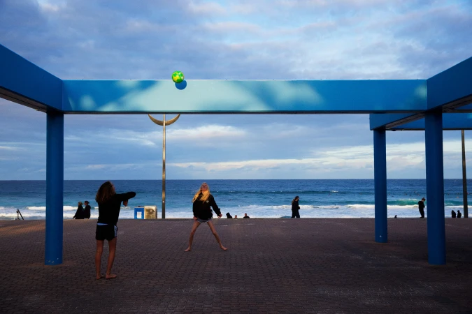 Two young women play with a soccer ball on a paved promenade by the sea under a blue-columned structure, beachgoers in background.