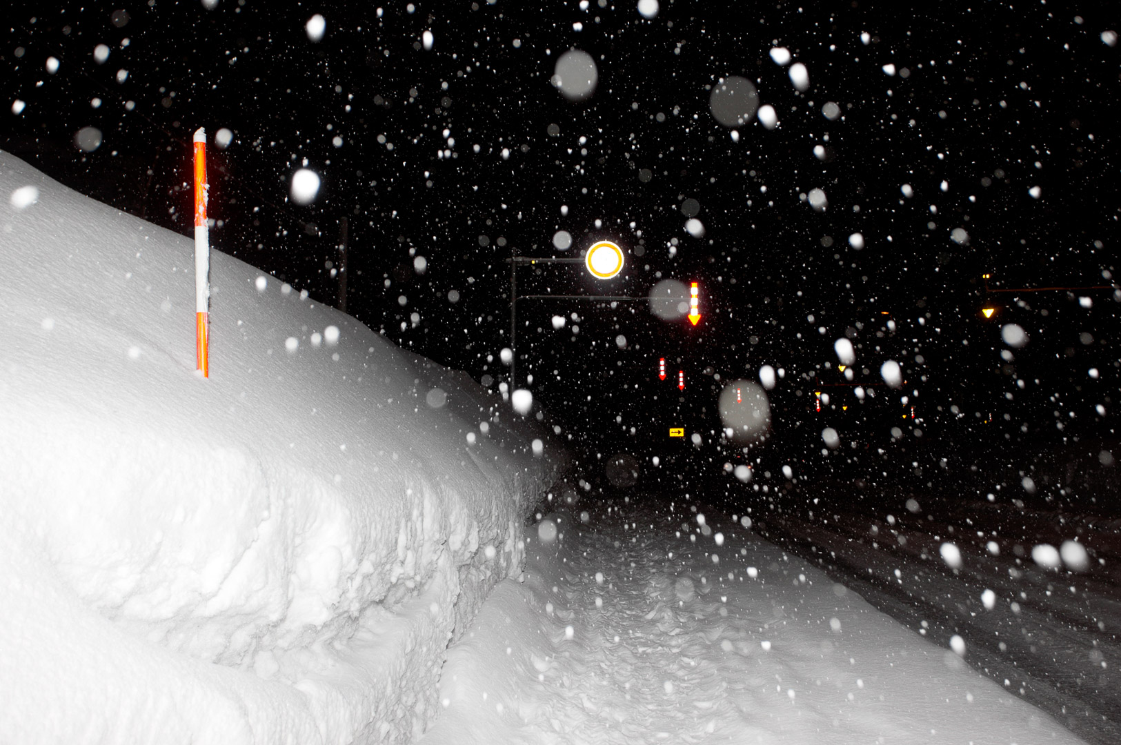 Snowy night scene with snow-covered roadside, a pole marker with orange bands, and streetlights in the distance under heavy snowfall.