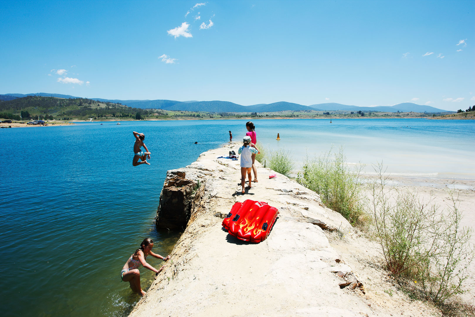 Children enjoy a summer day swimming and jumping off a rocky pier into a blue lake under a clear blue sky with distant mountains.