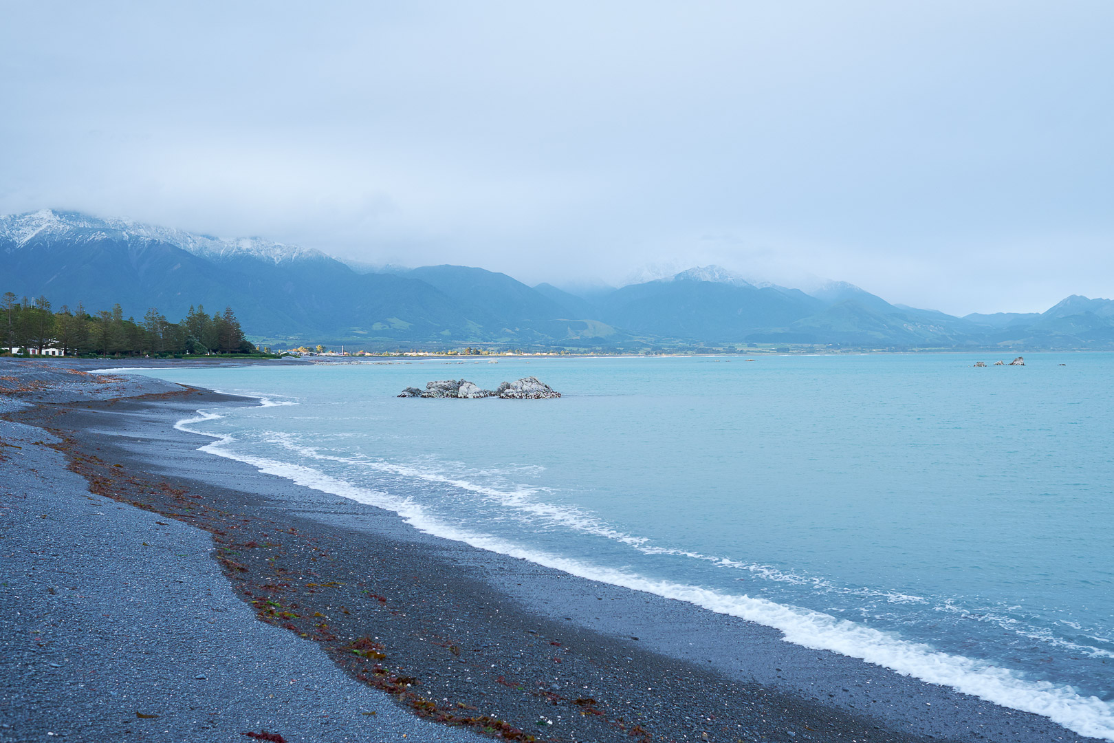 Scenic view of a rocky beach with waves, turquoise ocean, and snow-capped mountains under a cloudy sky on the horizon.