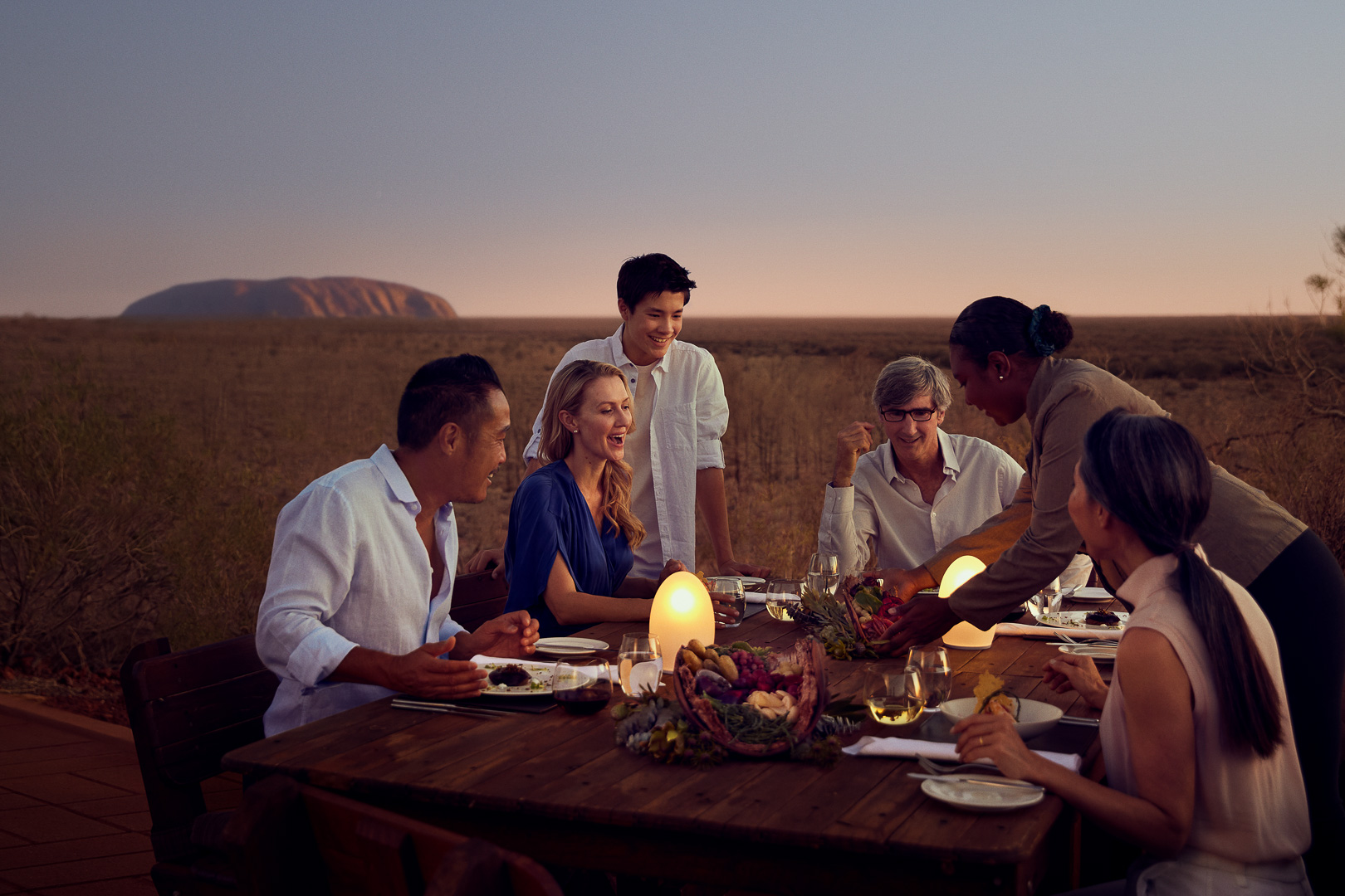 Group of people at an outdoor dining table enjoying a meal against the backdrop of a vast, dry landscape at dusk.