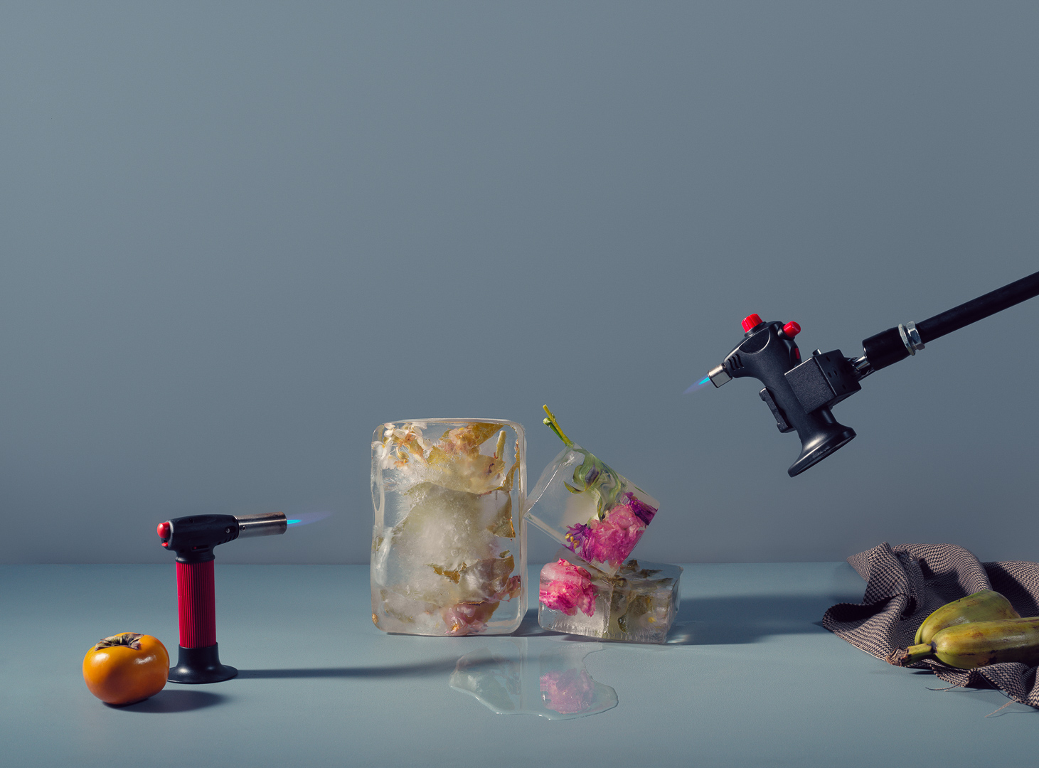 Still life of ice cubes with flowers inside, a persimmon, zucchini and two butane torches on a neutral blue-grey background.