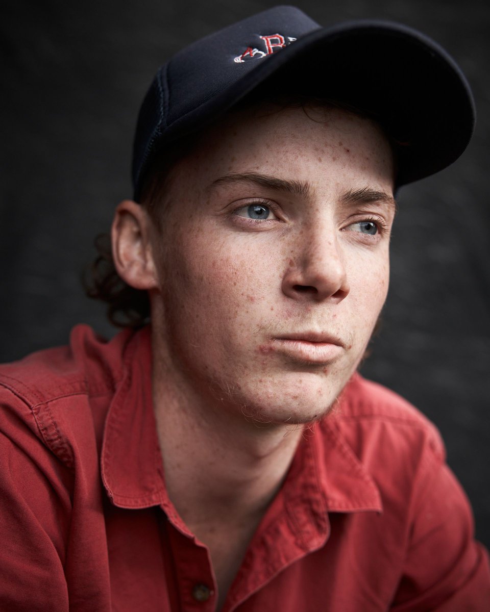 Portrait of a young adult male wearing a baseball cap and red button-down shirt, looking off to the side against a dark background.
