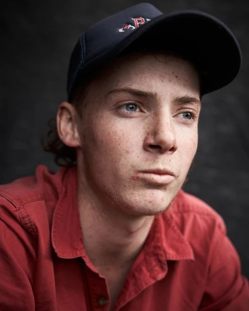 Portrait of a young adult male wearing a baseball cap and red button-down shirt, looking off to the side against a dark background.