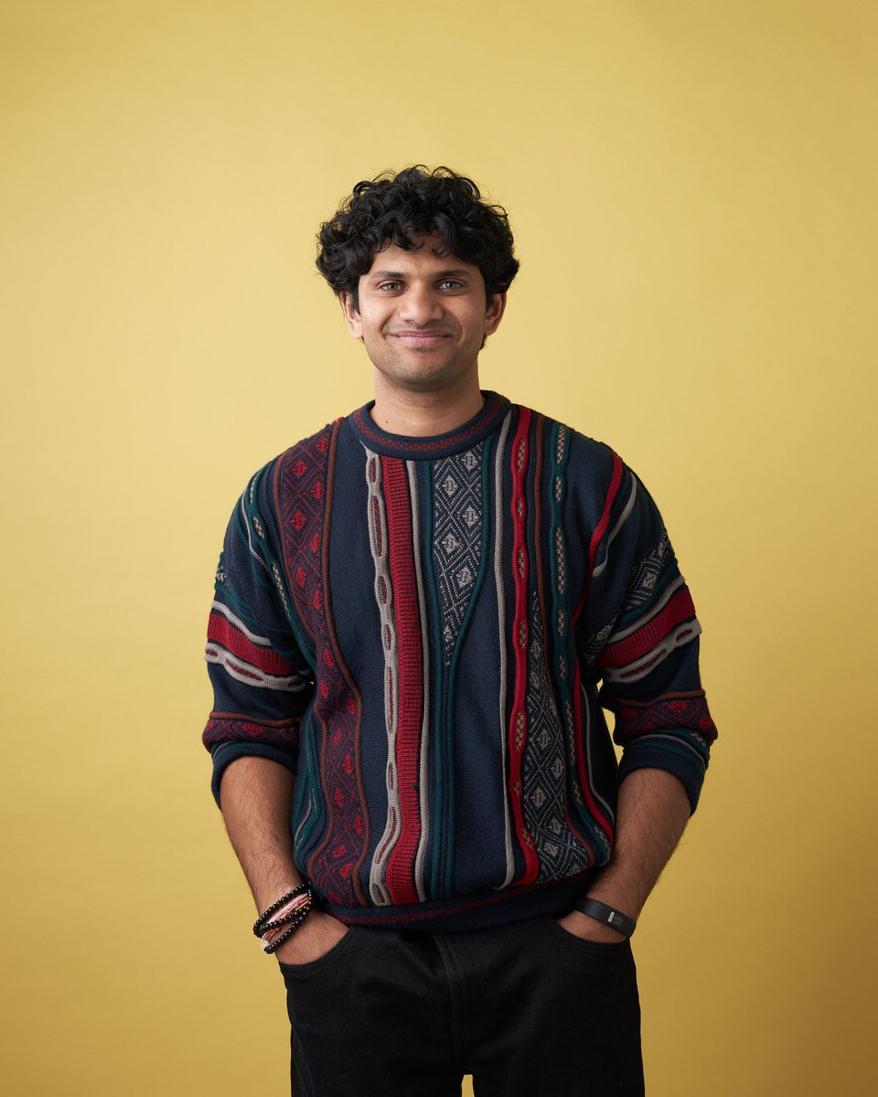 Portrait of a smiling dark-haired man wearing a patterned sweater against a solid yellow background, hands in his pockets, eye-level.