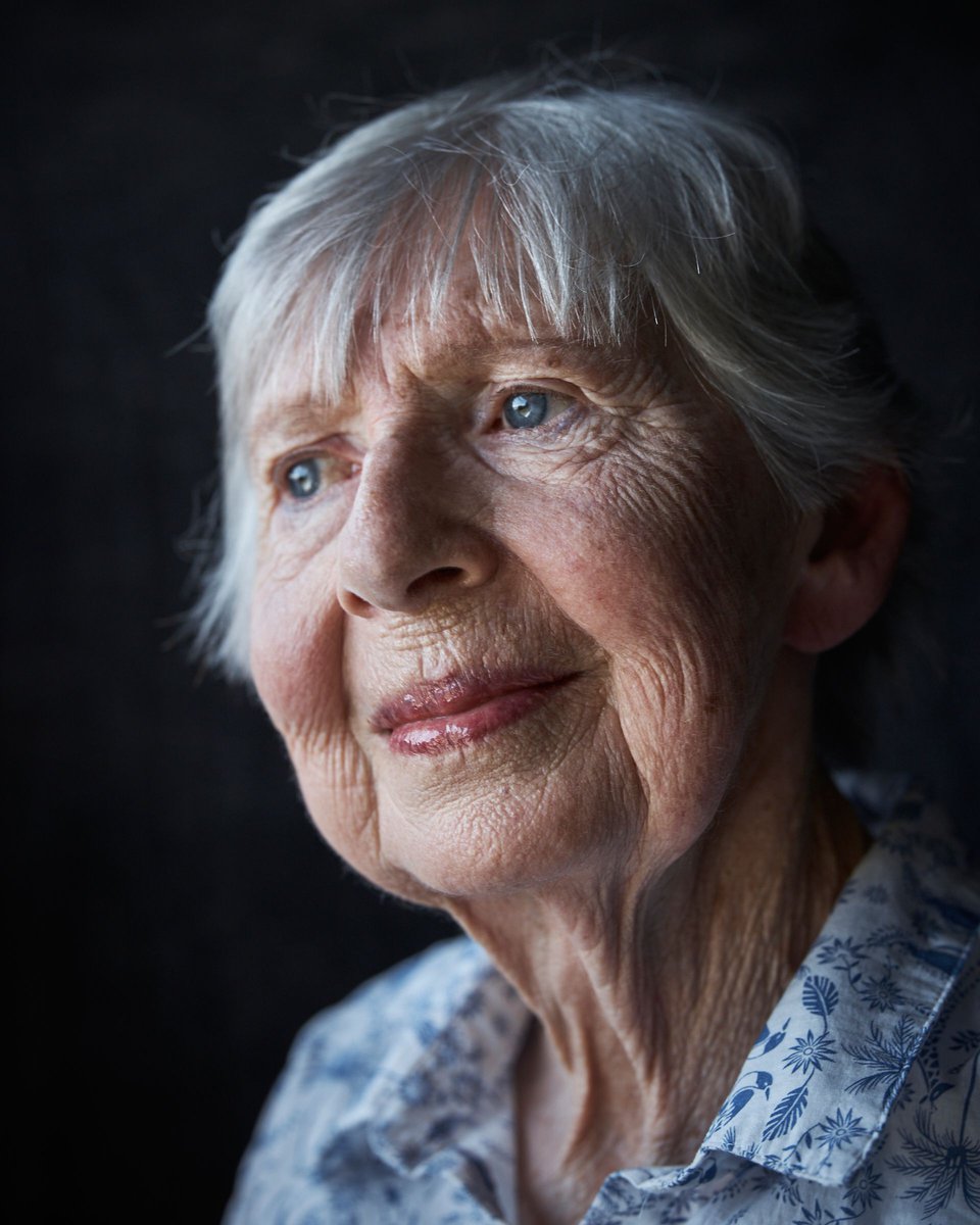 Close-up portrait of an elderly woman with gray hair, blue eyes, and a floral-patterned top against a dark background, looking up and to the left.