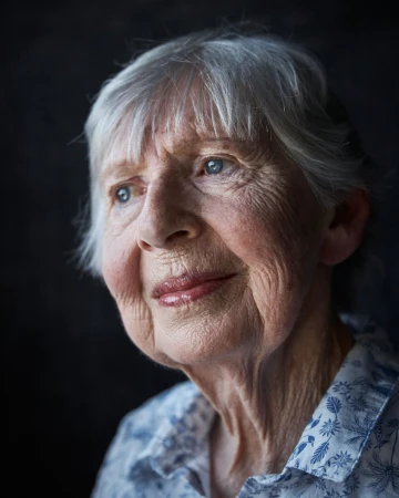 Close up portrait of an elderly woman with silver hair and blue eyes wearing a patterned shirt against a dark textured background