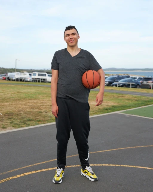 Full body shot of a young man holding a basketball on an outdoor court wearing athletic apparel, with cars parked near the ocean.