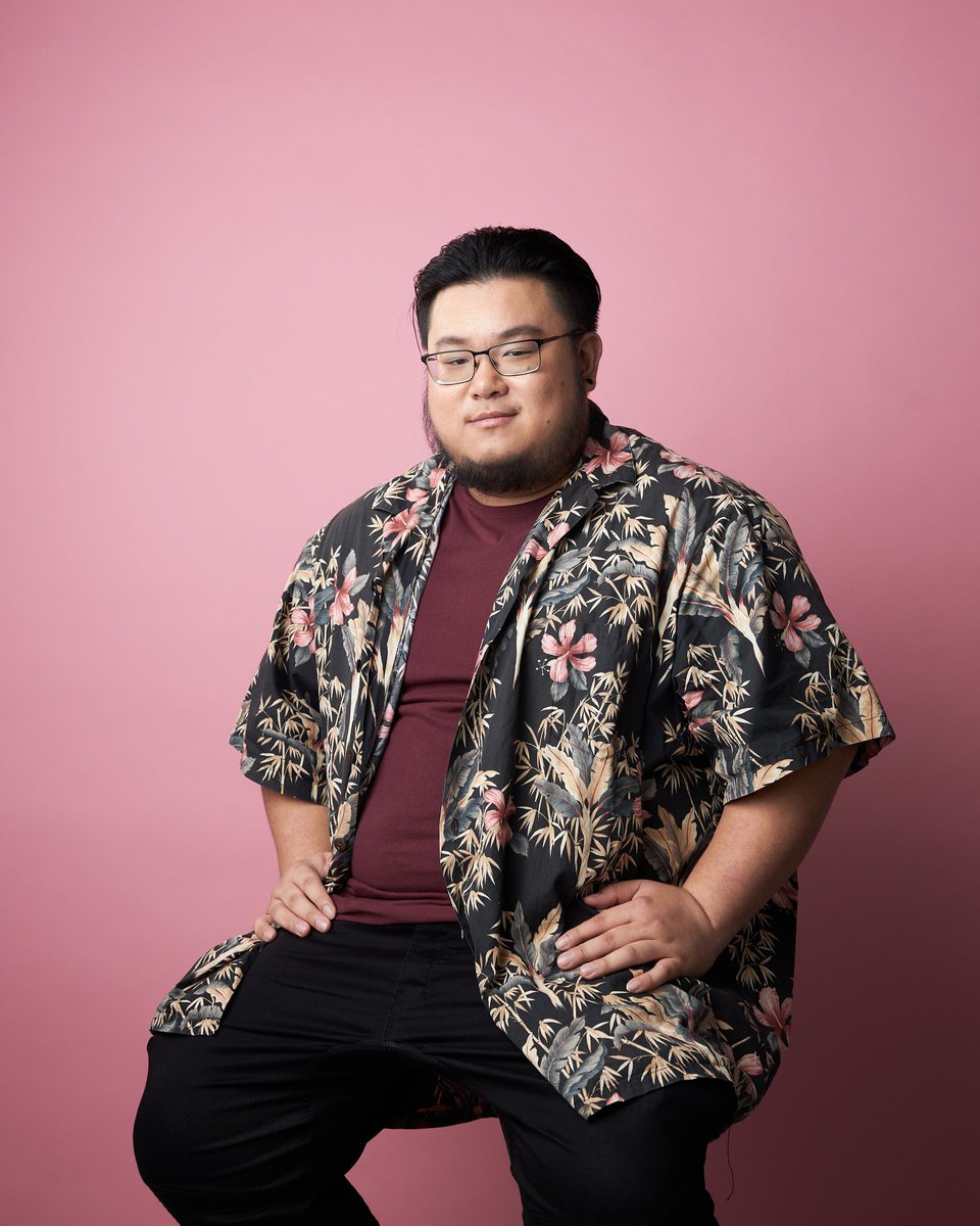 Portrait of an Asian man wearing glasses, a floral shirt, and black pants, sitting with his hands on his lap against a pink backdrop.