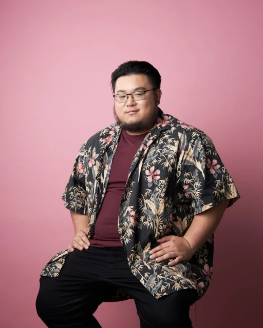 Portrait of an Asian man wearing glasses, a floral shirt, and black pants, sitting with his hands on his lap against a pink backdrop.