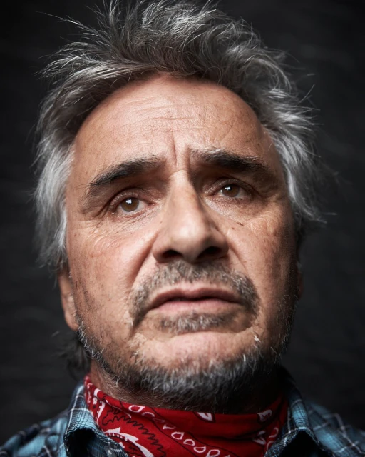 Portrait of an older man with salt and pepper hair and a red bandana around his neck, captured in a close-up shot against a dark background.