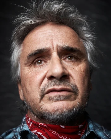 Portrait of an older man with salt and pepper hair and a red bandana around his neck, captured in a close-up shot against a dark background.