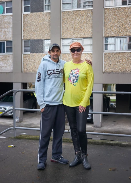 Two adults stand close together outdoors near an apartment building; the woman is wearing a yellow shirt and the man is wearing a light blue hoodie.