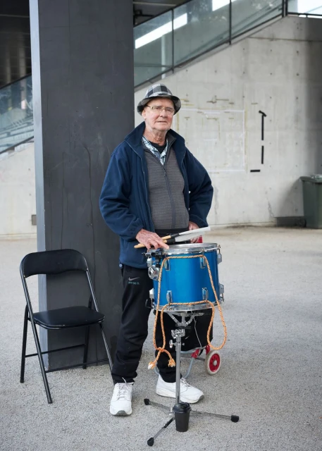 Elderly man plays a blue drum outdoors, wearing a hat and jacket; a black folding chair sits nearby in an urban setting.