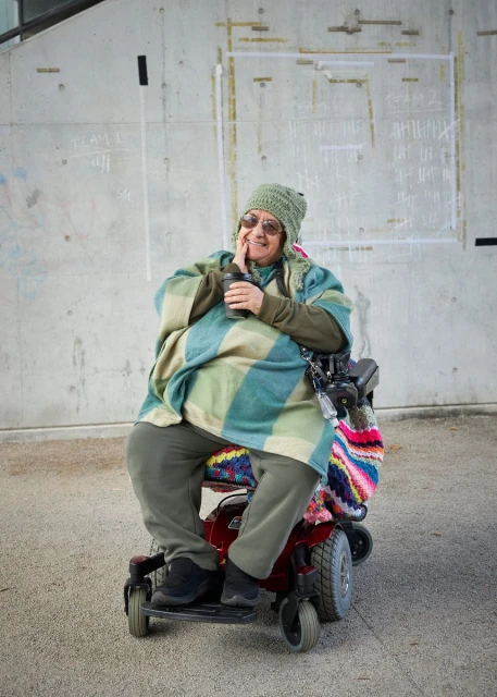 Adult woman with a patterned hat sits in a red electric wheelchair holding a cup in front of a weathered concrete wall outdoors.