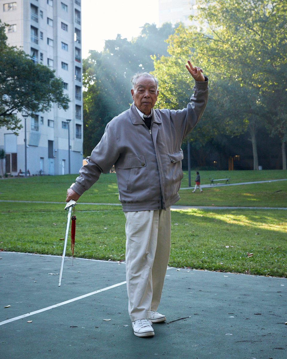 Elderly Asian man practices Tai Chi with a sword in a park with apartment buildings in the background, holding up two fingers.