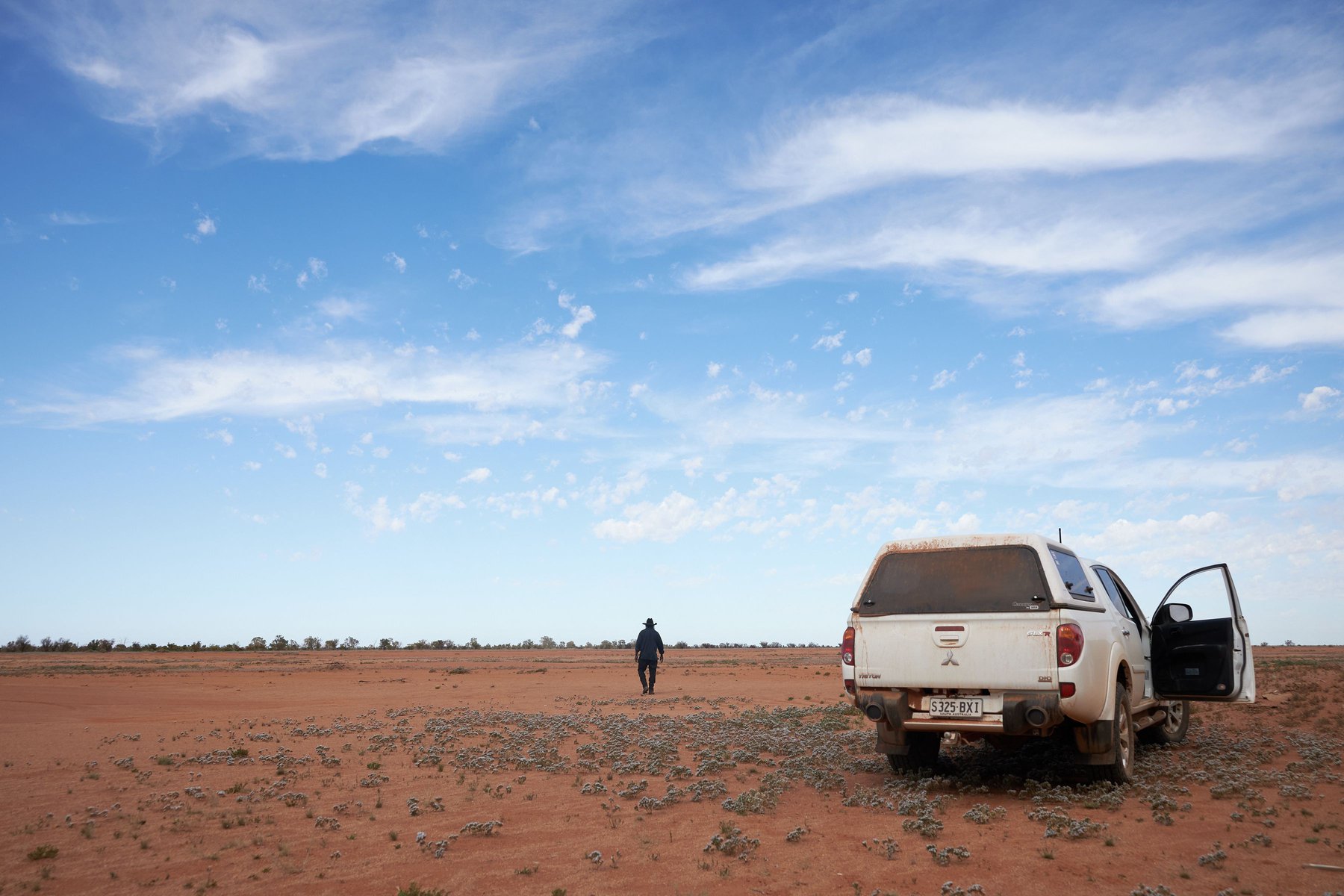 White pickup truck parked on a rural dirt landscape under a cloudy blue sky with a lone adult walking in the distance.