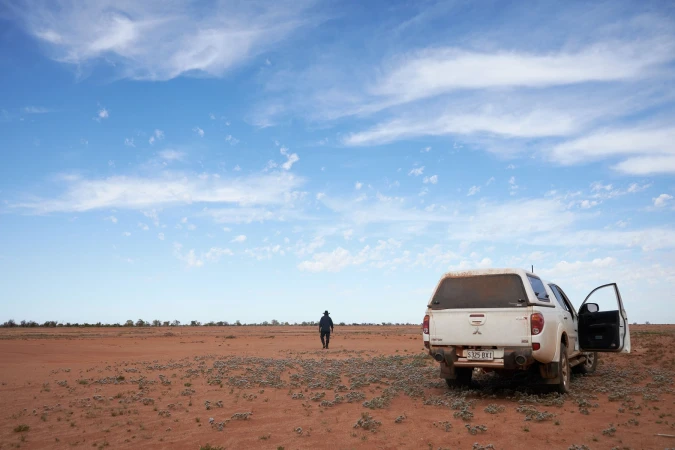 White pickup truck parked on a rural dirt landscape under a cloudy blue sky with a lone adult walking in the distance. 