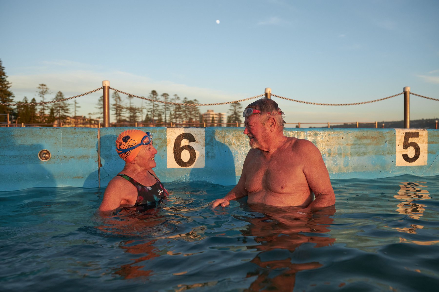 Older couple in swimwear and goggles standing in a pool; concrete pool edge with numbers; sky, trees, and buildings visible in the background.