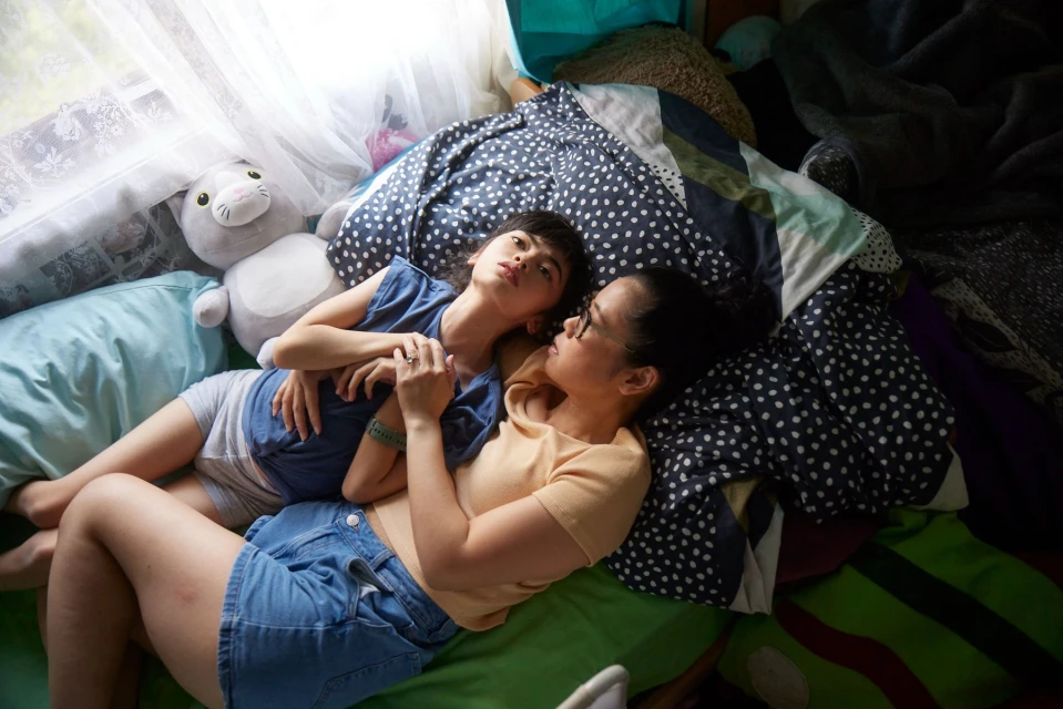 Mother and child lay on a bed with patterned bedding and pillows near a sunlit window in a cozy, intimate indoor setting.