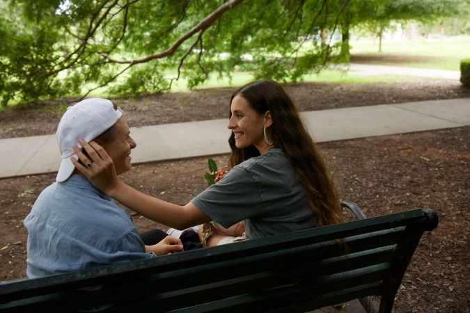 A smiling couple sitting together on a dark park bench outdoors beneath leafy trees, captured in profile by photographer Joel Pratley.