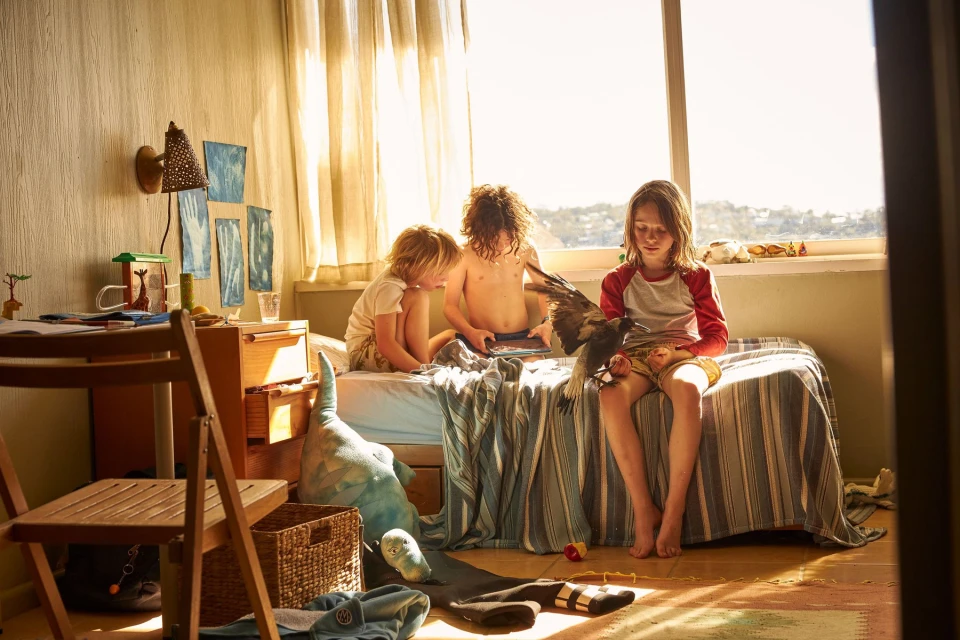 Three children are sitting on a bed in a brightly lit room, one holding a bird with spread wings, with a window in the background.