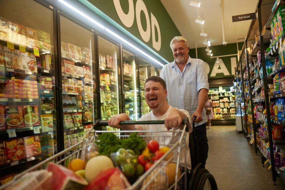 Two men in a grocery store: one in a wheelchair laughing, the other pushing the cart filled with colorful fruits and vegetables.