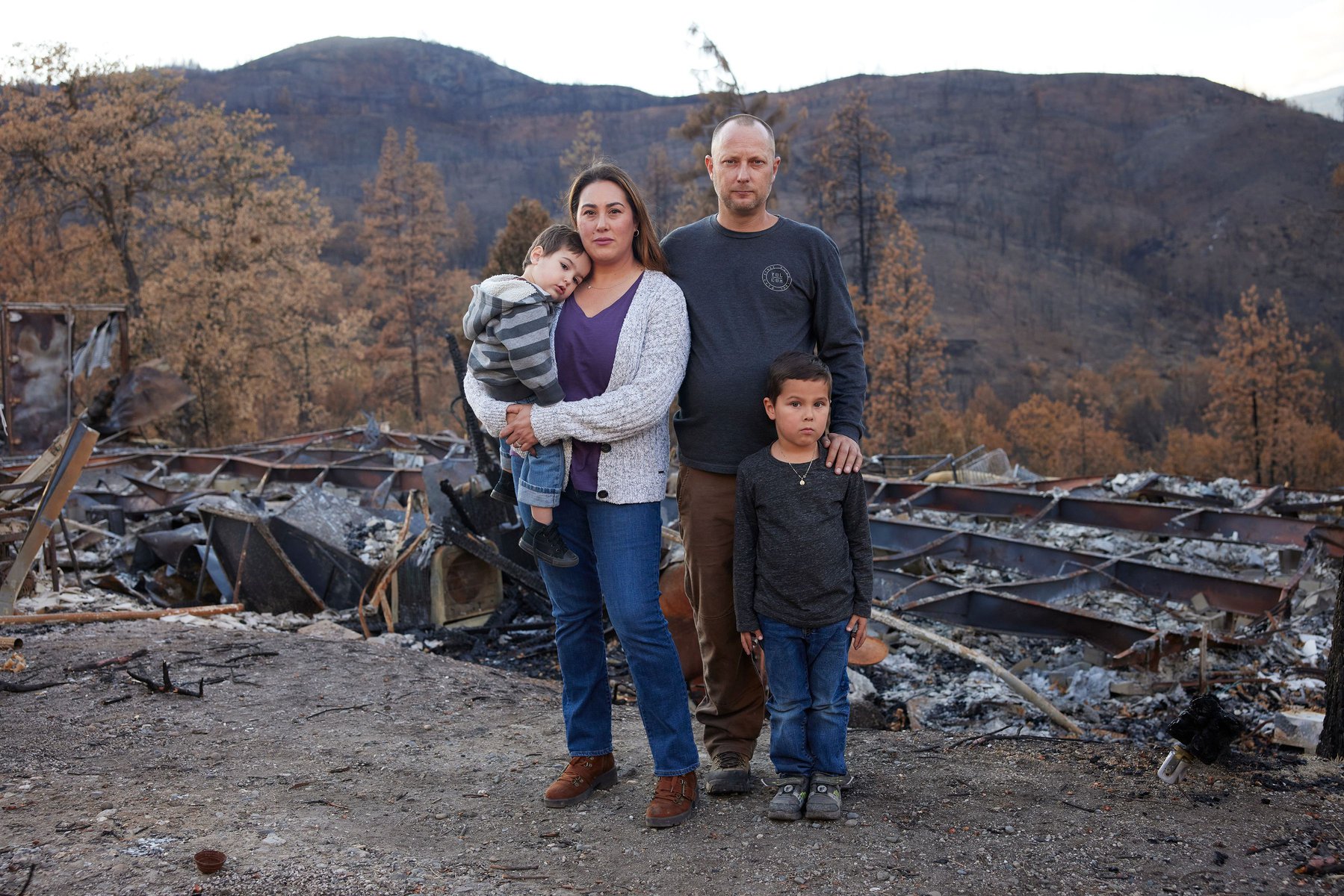 Family of four stands amidst the aftermath of a fire, with a destroyed structure and scorched landscape visible behind them.