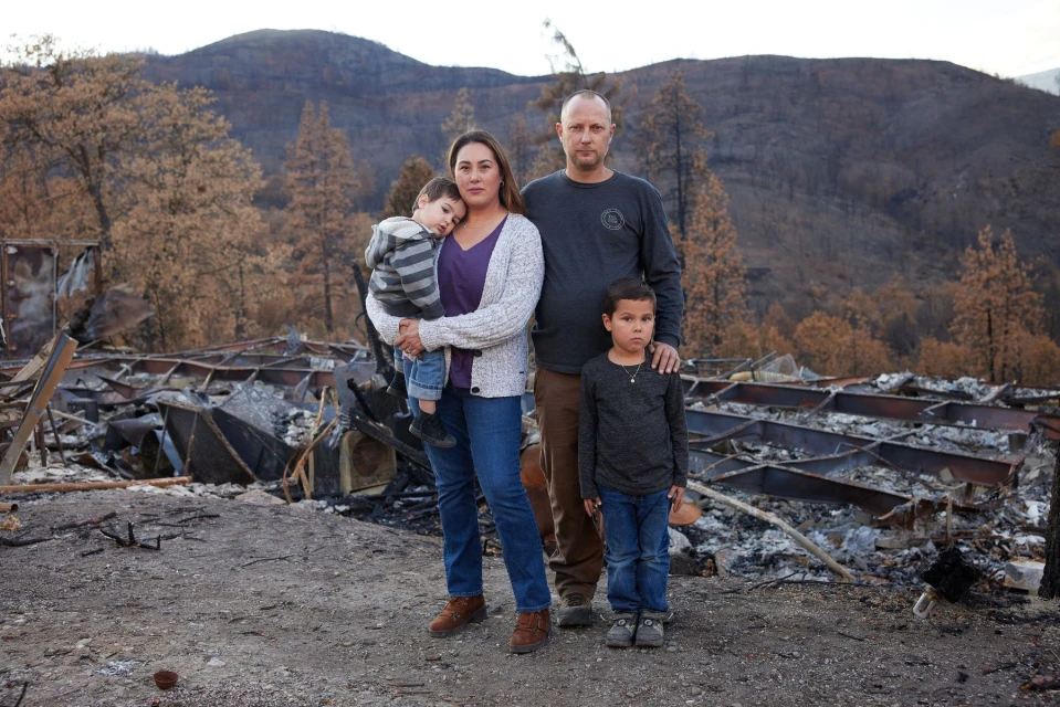 Family of four stands amidst the aftermath of a fire, with a destroyed structure and scorched landscape visible behind them.