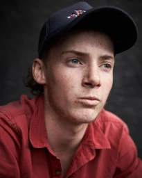 Close-up portrait of a young man wearing a red shirt and a dark baseball cap against a dark textured background.