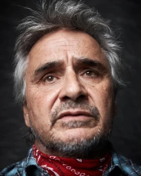 Close-up portrait of an older man with grey hair wearing a plaid shirt and red neckerchief against a dark textured background.