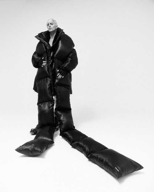 Fashionable person in dramatic black puffer coat with long padded appendages against a white background in a studio setting.