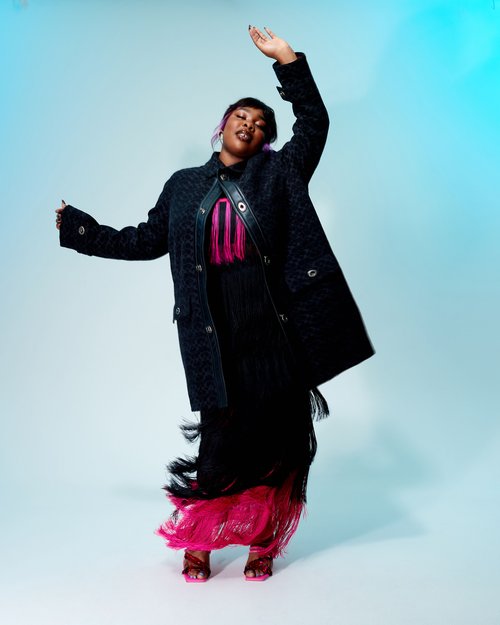 A fashionable Black woman with dyed hair poses in an outwear coat with fringe detail on a graduated blue background in a studio shot.
