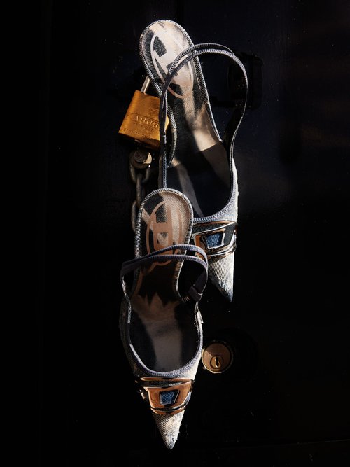 Pointed-toe slingback heels with gold accents, secured by a padlock and chain, against a dark background in a high-fashion still life.