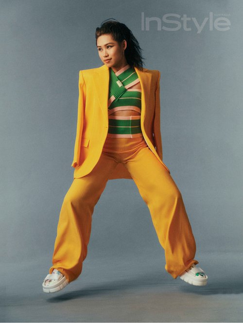 An Asian female fashion model is jumping and posing in a yellow suit and sneakers on a grey background in a studio fashion shot.