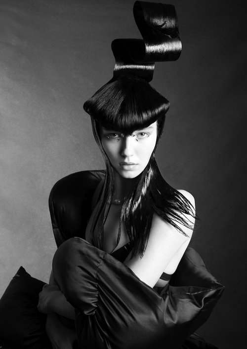 Monochrome portrait of a woman with stylized black hair, wearing a black top and dark pants, seated against a grey background.