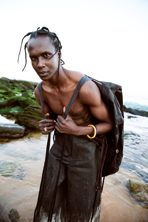 Adult male with dreadlocks and bare chest poses with a backpack on a rocky beach with green rocks and clear water in an eye-level shot.