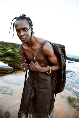 Adult male with dreadlocks and bare chest poses with a backpack on a rocky beach with green rocks and clear water in an eye-level shot.