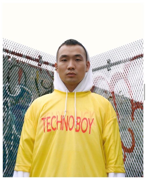 Portrait of an Asian man wearing a bright yellow hoodie with white hood, in front of graffiti on a chain-link fence.