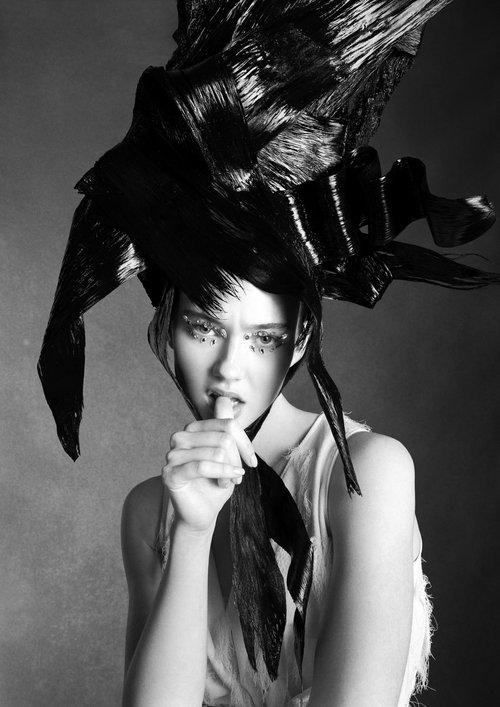 Black and white portrait of a woman with dramatic black hair and embellished makeup, in a sleeveless top against a grey backdrop.