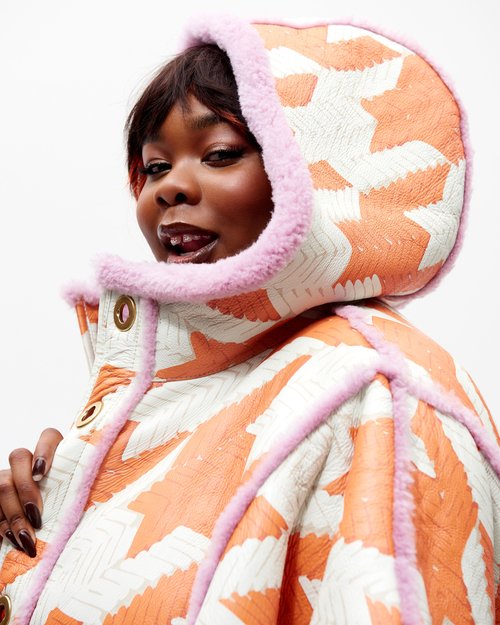 Portrait of a Black woman wearing an orange and white patterned jacket with pink fur trim against a white background.