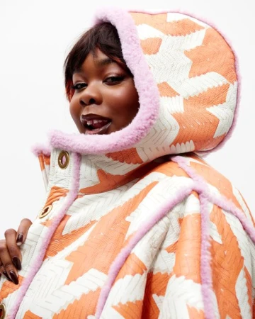 Portrait of a Black woman wearing an orange and white patterned jacket with pink fur trim against a white background.