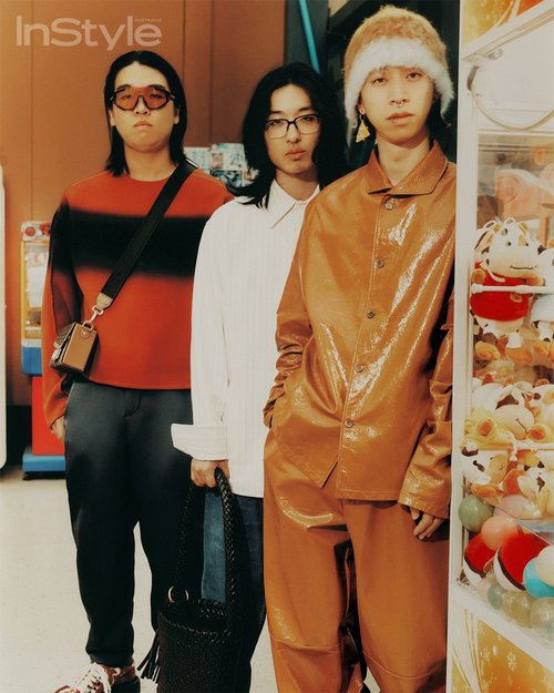 Three stylish individuals pose indoors: one in a striped top, another in white, and one in orange, by a claw machine.