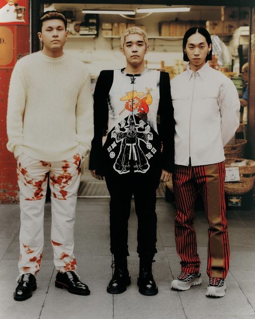 Three adult males pose in front of a food store, displaying unique fashion ensembles with neutral colors and patterned pants.